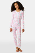A young girl with curly hair stands barefoot on a plain background, wearing Roller Rabbit’s Kids Sleepy Sloth Pajamas in pink and white with playful animal prints.
