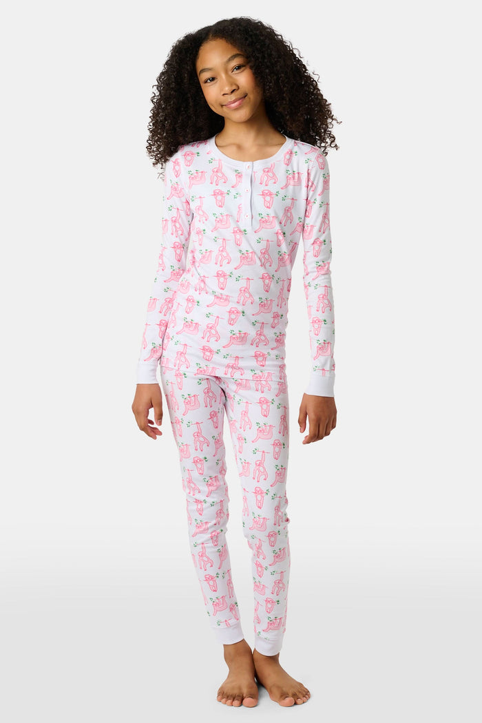 A young girl with curly hair stands barefoot on a plain background, wearing Roller Rabbit’s Kids Sleepy Sloth Pajamas in pink and white with playful animal prints.