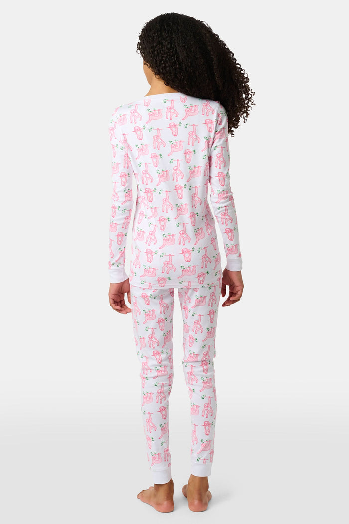 A young girl with curly hair stands barefoot on a plain background, wearing Roller Rabbit’s Kids Sleepy Sloth Pajamas in pink and white with playful animal prints.