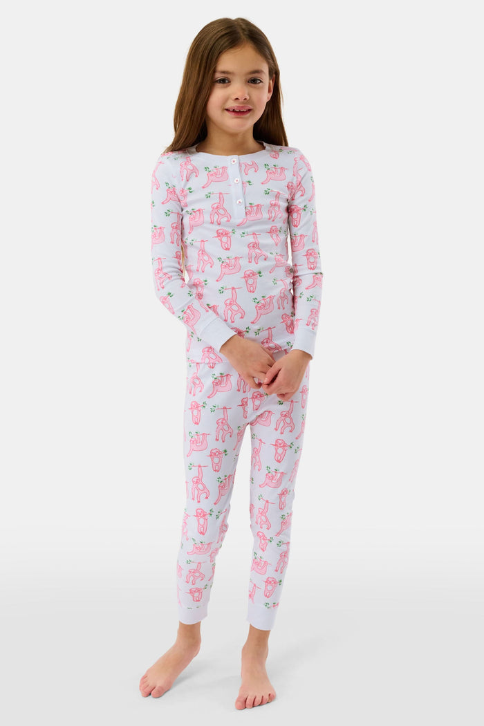 A young girl with curly hair stands barefoot on a plain background, wearing Roller Rabbit’s Kids Sleepy Sloth Pajamas in pink and white with playful animal prints.