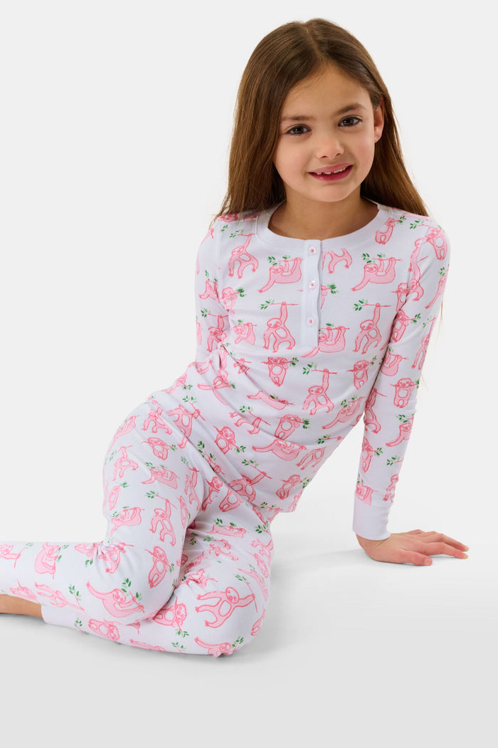 A young girl with curly hair stands barefoot on a plain background, wearing Roller Rabbit’s Kids Sleepy Sloth Pajamas in pink and white with playful animal prints.