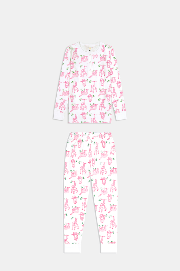 A young girl with curly hair stands barefoot on a plain background, wearing Roller Rabbit’s Kids Sleepy Sloth Pajamas in pink and white with playful animal prints.