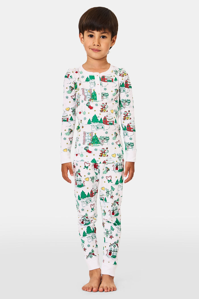 A young child stands barefoot in Roller Rabbit Kids Snow Village Pajamas—white, long-sleeve Pima cotton decorated with colorful Christmas trees, gifts, candy canes, and festive scenes—against a plain white background.