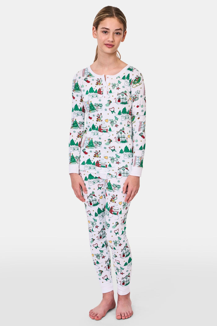 A young child stands barefoot in Roller Rabbit Kids Snow Village Pajamas—white, long-sleeve Pima cotton decorated with colorful Christmas trees, gifts, candy canes, and festive scenes—against a plain white background.