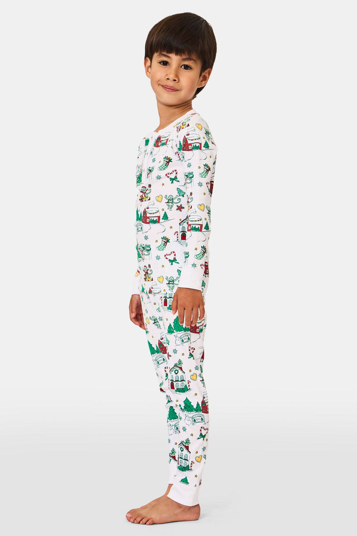 A young child stands barefoot in Roller Rabbit Kids Snow Village Pajamas—white, long-sleeve Pima cotton decorated with colorful Christmas trees, gifts, candy canes, and festive scenes—against a plain white background.