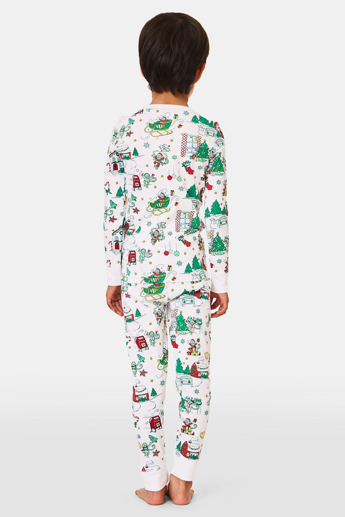 A young child stands barefoot in Roller Rabbit Kids Snow Village Pajamas—white, long-sleeve Pima cotton decorated with colorful Christmas trees, gifts, candy canes, and festive scenes—against a plain white background.