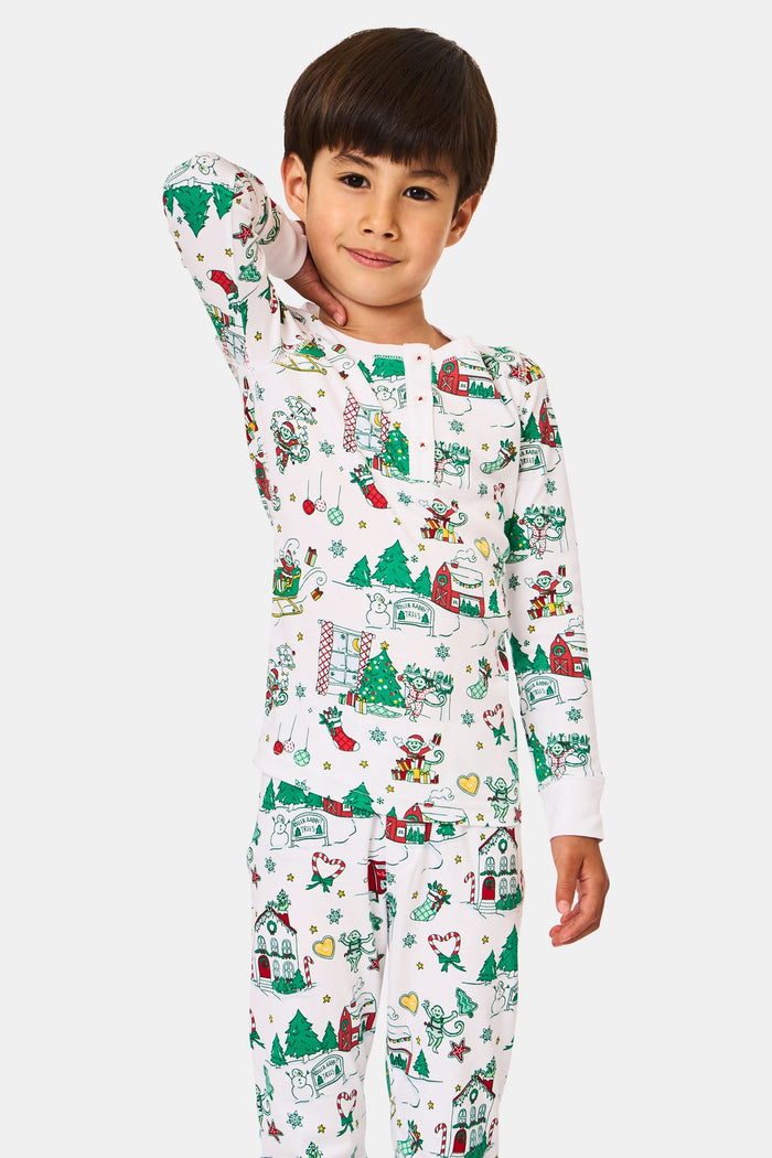 A young child stands barefoot in Roller Rabbit Kids Snow Village Pajamas—white, long-sleeve Pima cotton decorated with colorful Christmas trees, gifts, candy canes, and festive scenes—against a plain white background.