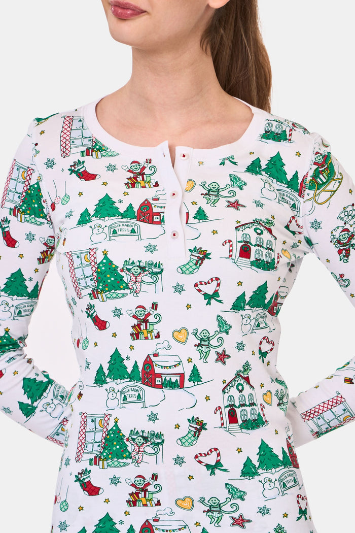 A young child stands barefoot in Roller Rabbit Kids Snow Village Pajamas—white, long-sleeve Pima cotton decorated with colorful Christmas trees, gifts, candy canes, and festive scenes—against a plain white background.