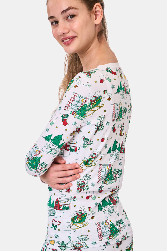 A young child stands barefoot in Roller Rabbit Kids Snow Village Pajamas—white, long-sleeve Pima cotton decorated with colorful Christmas trees, gifts, candy canes, and festive scenes—against a plain white background.