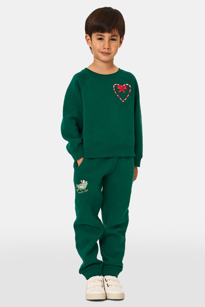 A young child smiles, wearing Roller Rabbit Kids Snow Village Sweatpants—matching green cotton joggers with embroidered designs—and a sweatshirt with white sneakers, against a plain light background.