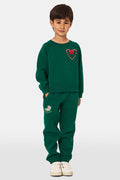 A young child smiles, wearing Roller Rabbit Kids Snow Village Sweatpants—matching green cotton joggers with embroidered designs—and a sweatshirt with white sneakers, against a plain light background.