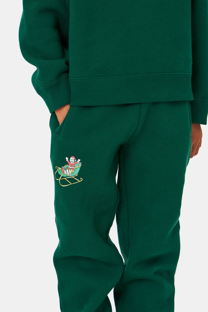 A young child smiles, wearing Roller Rabbit Kids Snow Village Sweatpants—matching green cotton joggers with embroidered designs—and a sweatshirt with white sneakers, against a plain light background.