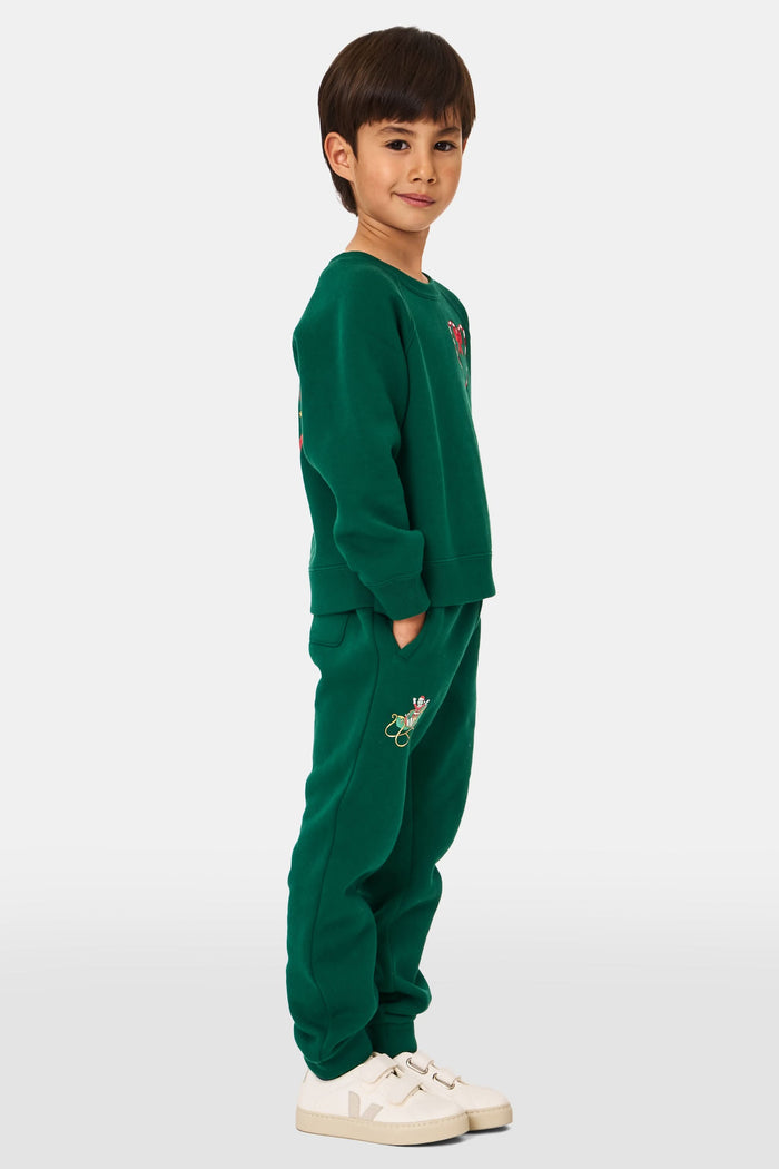 A young child smiles, wearing Roller Rabbit Kids Snow Village Sweatpants—matching green cotton joggers with embroidered designs—and a sweatshirt with white sneakers, against a plain light background.