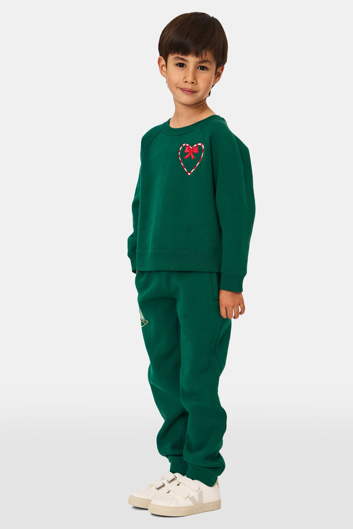 A young child smiles, wearing Roller Rabbit Kids Snow Village Sweatpants—matching green cotton joggers with embroidered designs—and a sweatshirt with white sneakers, against a plain light background.