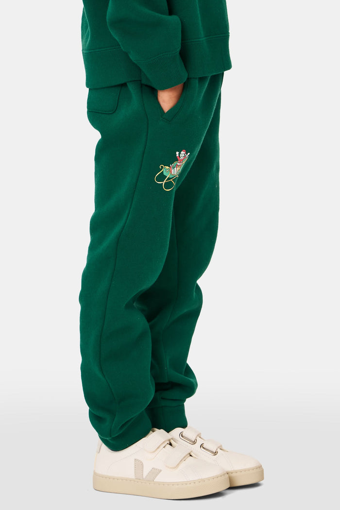 A young child smiles, wearing Roller Rabbit Kids Snow Village Sweatpants—matching green cotton joggers with embroidered designs—and a sweatshirt with white sneakers, against a plain light background.