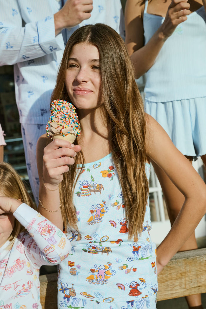Young child with long brown hair stands barefoot, wearing the Roller Rabbit Kids Spain Monkey Noa Set—light blue Pima cotton loungewear with playful monkey prints and blue trim—against a plain white background.