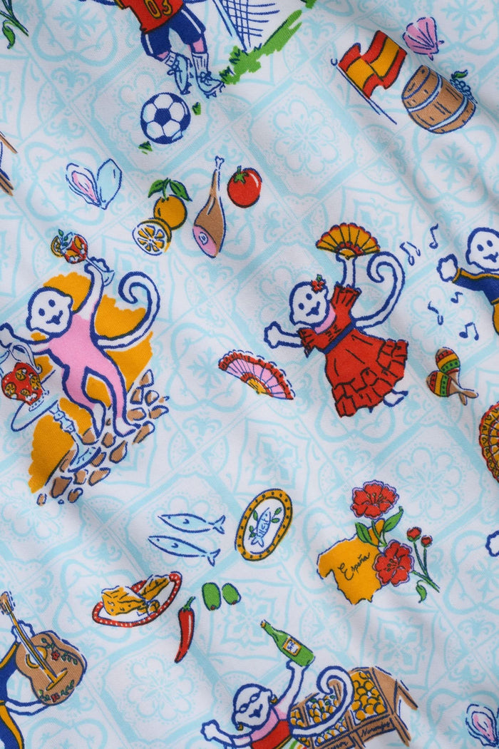 A woman with curly hair smiles in Roller Rabbit Spain Monkey Polo Pajamas—light blue shortie PJs made from 100 Pima cotton with colorful whimsical prints and red piping—standing against a plain white background.