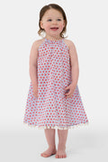 A young girl with long brown hair stands facing forward in the Roller Rabbit Kids Starry Dot Carrie Cover Up—a sleeveless, knee-length white dress featuring a red dotted pattern and pom-pom trim. She has a neutral expression and a light background behind her.