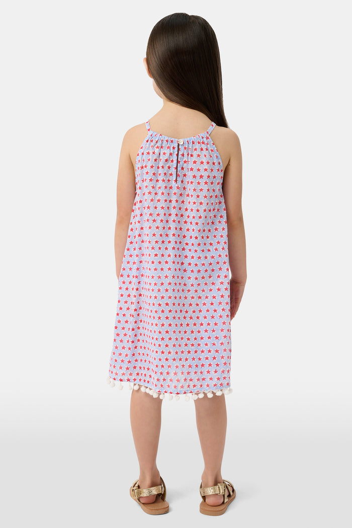 A young girl with long brown hair stands facing forward in the Roller Rabbit Kids Starry Dot Carrie Cover Up—a sleeveless, knee-length white dress featuring a red dotted pattern and pom-pom trim. She has a neutral expression and a light background behind her.