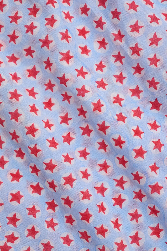 A young girl with long brown hair stands facing forward in the Roller Rabbit Kids Starry Dot Carrie Cover Up—a sleeveless, knee-length white dress featuring a red dotted pattern and pom-pom trim. She has a neutral expression and a light background behind her.