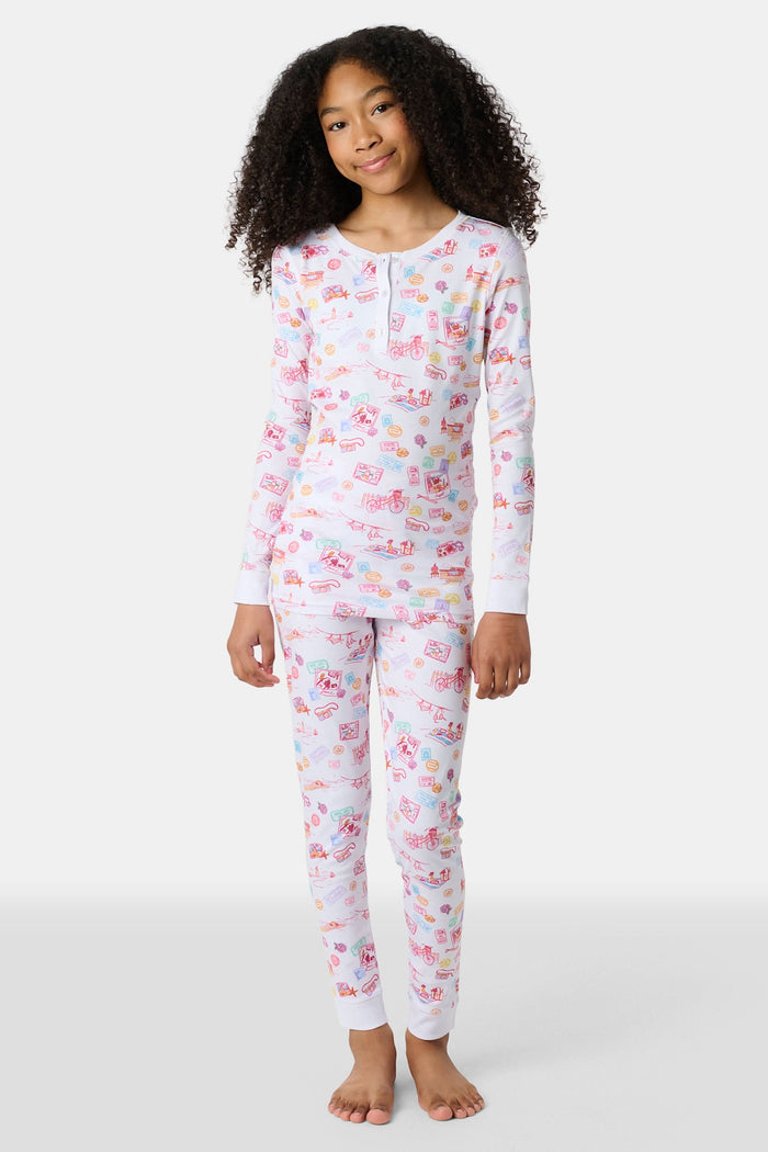A young girl smiles barefoot in Roller Rabbit Kids Summer Memories Pajamas, made from soft Pima Cotton with playful, colorful prints on white. She stands against a light gray background. Made in Peru.