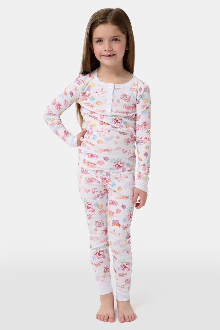 A young girl smiles barefoot in Roller Rabbit Kids Summer Memories Pajamas, made from soft Pima Cotton with playful, colorful prints on white. She stands against a light gray background. Made in Peru.