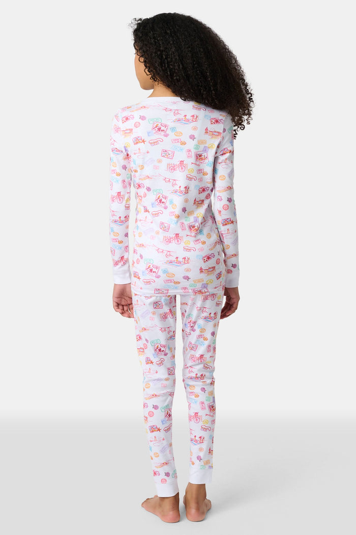 A young girl smiles barefoot in Roller Rabbit Kids Summer Memories Pajamas, made from soft Pima Cotton with playful, colorful prints on white. She stands against a light gray background. Made in Peru.