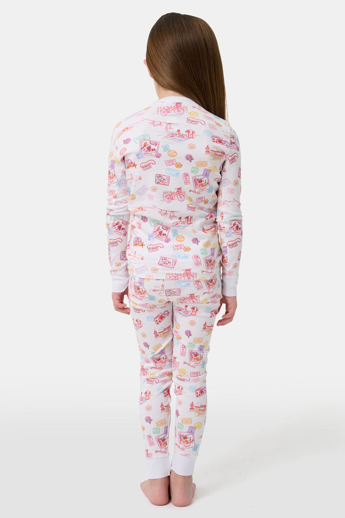 A young girl smiles barefoot in Roller Rabbit Kids Summer Memories Pajamas, made from soft Pima Cotton with playful, colorful prints on white. She stands against a light gray background. Made in Peru.