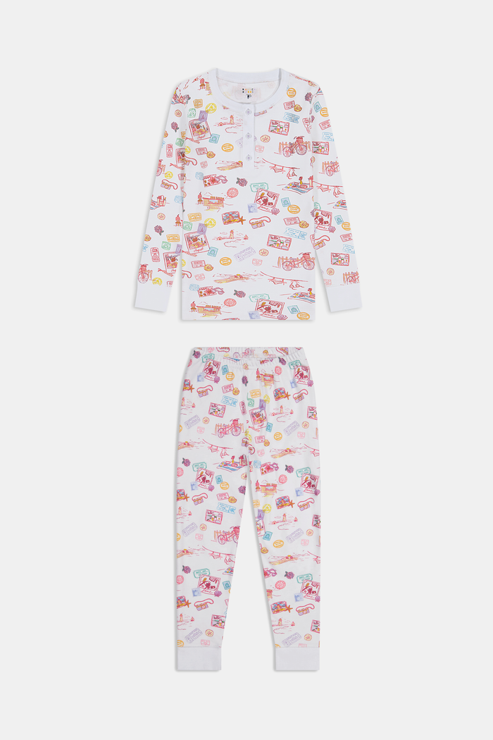 A young girl smiles barefoot in Roller Rabbit Kids Summer Memories Pajamas, made from soft Pima Cotton with playful, colorful prints on white. She stands against a light gray background. Made in Peru.