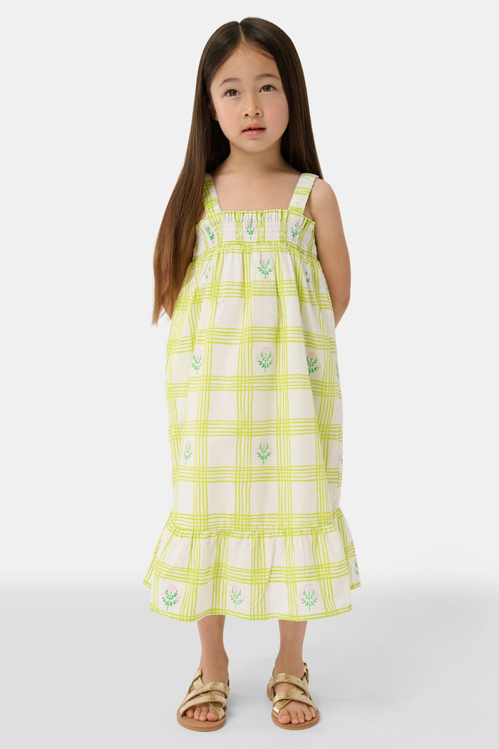 A young girl with long brown hair wears the Roller Rabbit Kids Sun Check Blossom Teagan Dress—a yellow-and-white plaid sundress with ruffled hem and green floral accents. She stands facing forward, hands behind her back, in gold sandals.
