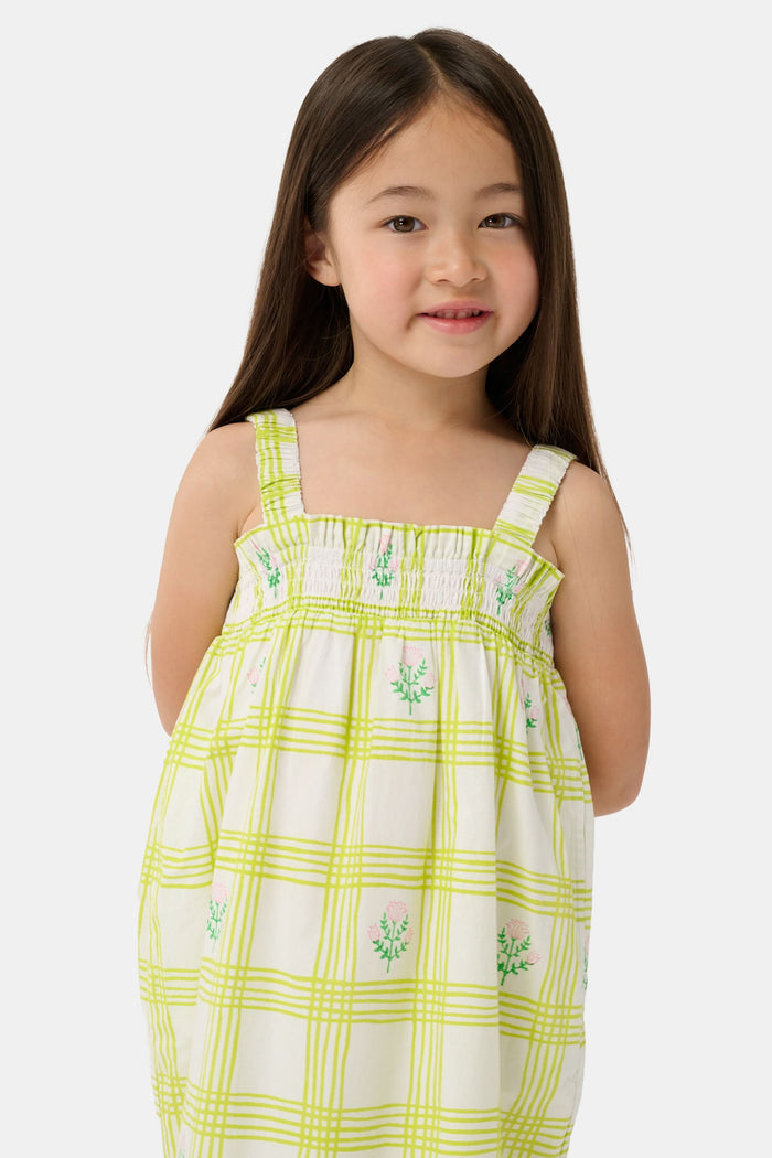 A young girl with long brown hair wears the Roller Rabbit Kids Sun Check Blossom Teagan Dress—a yellow-and-white plaid sundress with ruffled hem and green floral accents. She stands facing forward, hands behind her back, in gold sandals.