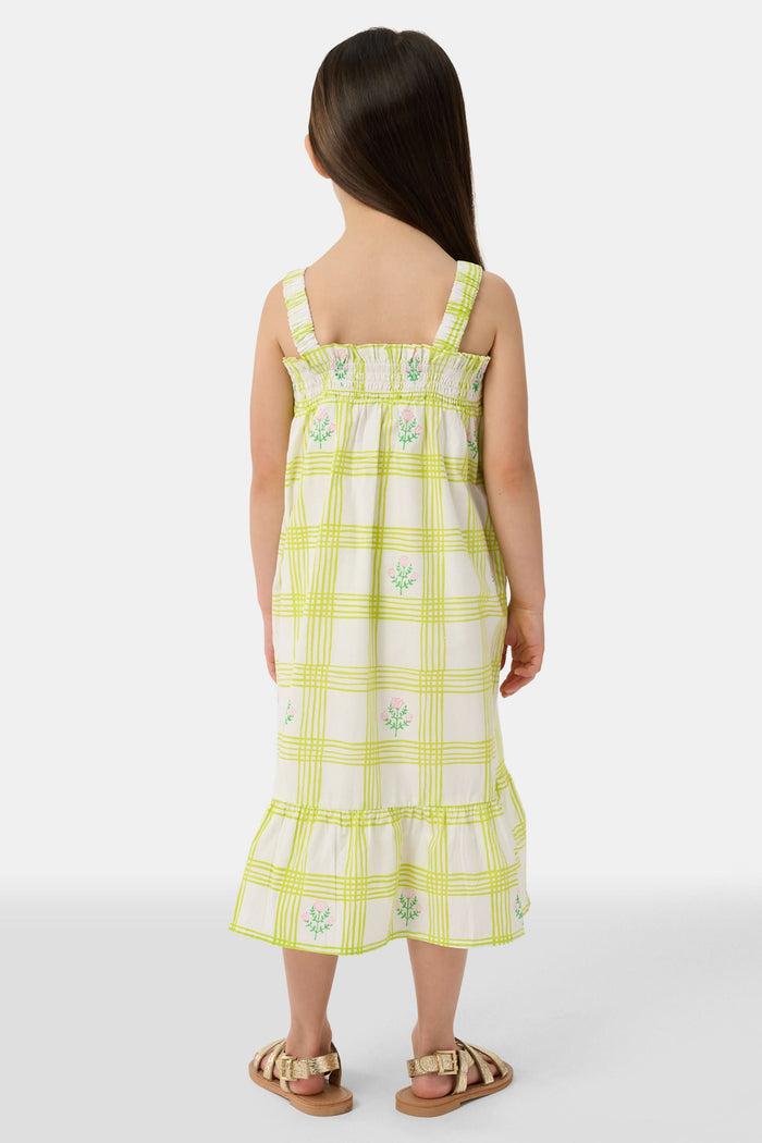 A young girl with long brown hair wears the Roller Rabbit Kids Sun Check Blossom Teagan Dress—a yellow-and-white plaid sundress with ruffled hem and green floral accents. She stands facing forward, hands behind her back, in gold sandals.
