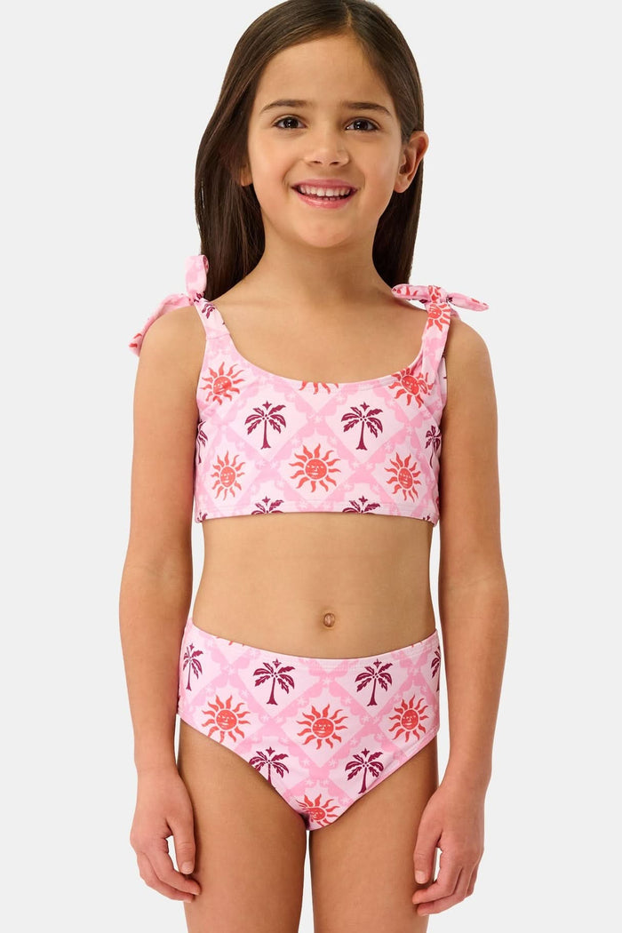 A young girl with long brown hair smiles in a Roller Rabbit Kids Sunny Days Cora Bikini, featuring sun and palm tree patterns and adjustable straps, against a plain light background.