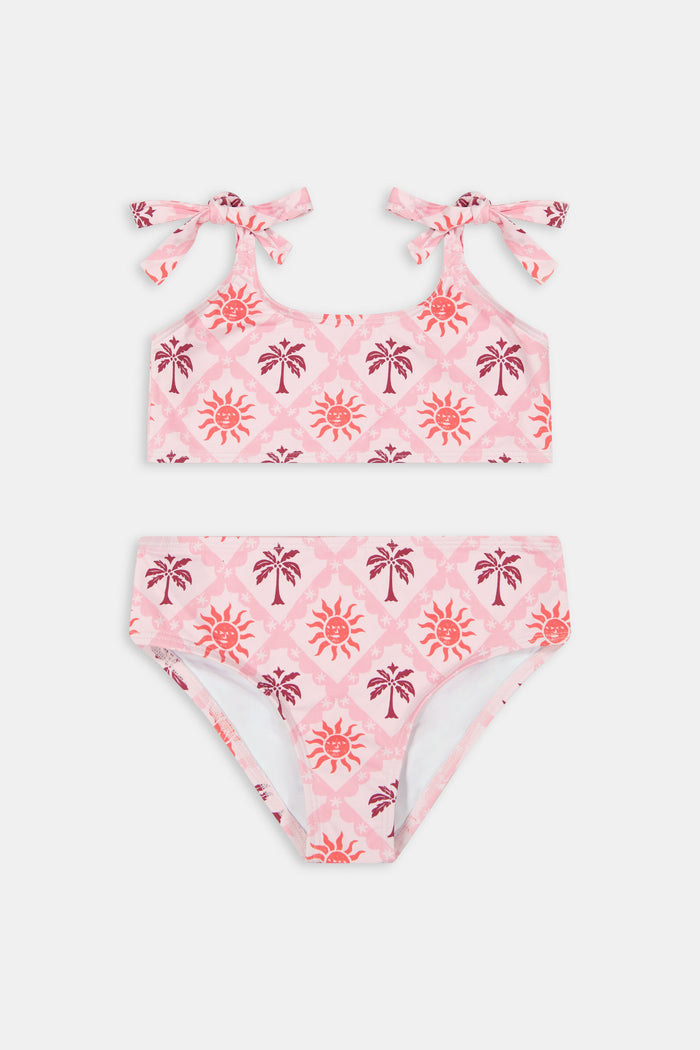 A young girl with long brown hair smiles in a Roller Rabbit Kids Sunny Days Cora Bikini, featuring sun and palm tree patterns and adjustable straps, against a plain light background.