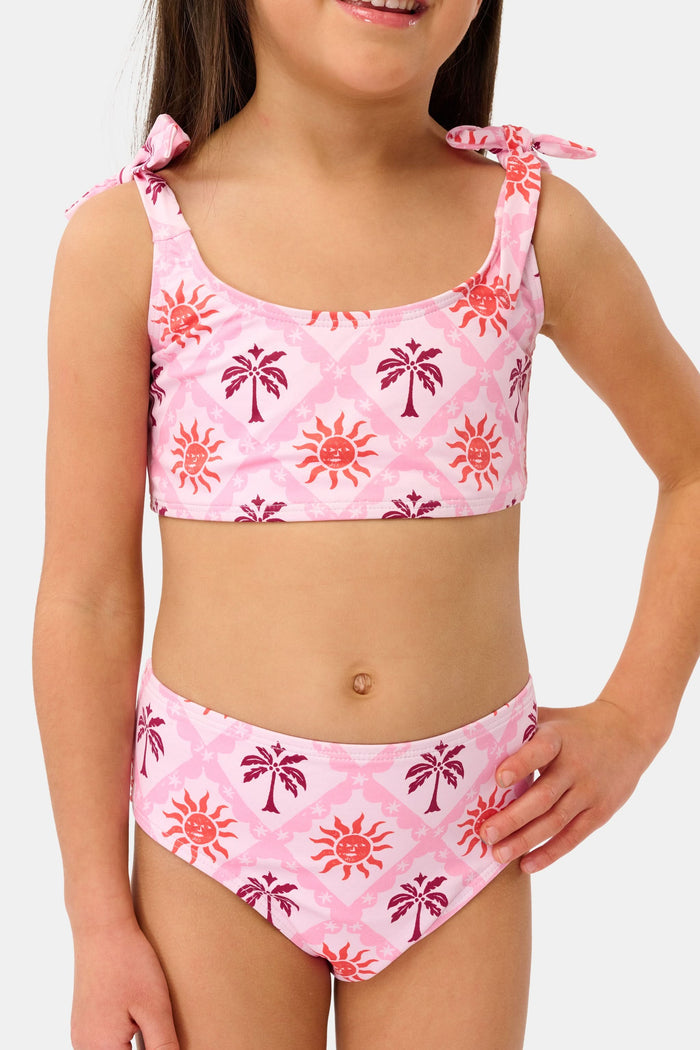 A young girl with long brown hair smiles in a Roller Rabbit Kids Sunny Days Cora Bikini, featuring sun and palm tree patterns and adjustable straps, against a plain light background.