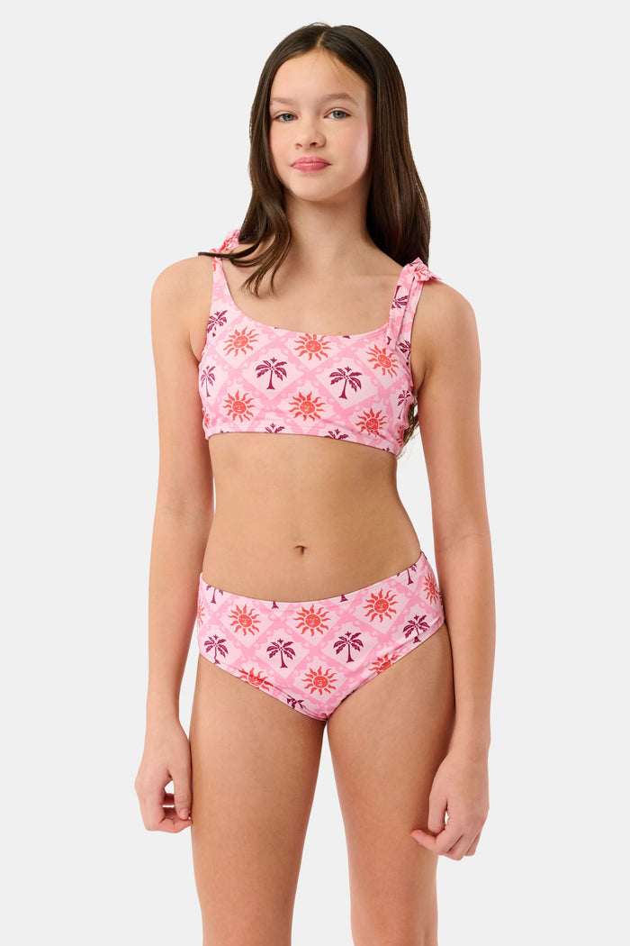 A young girl with long brown hair smiles in a Roller Rabbit Kids Sunny Days Cora Bikini, featuring sun and palm tree patterns and adjustable straps, against a plain light background.