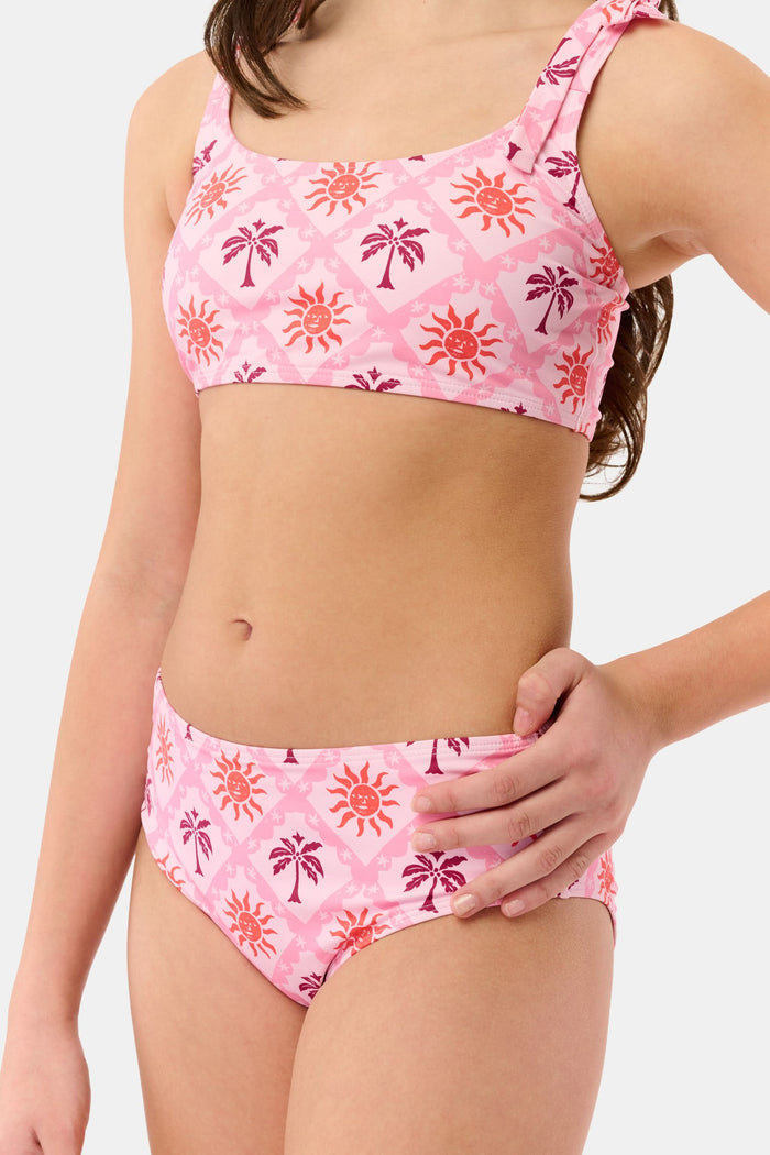 A young girl with long brown hair smiles in a Roller Rabbit Kids Sunny Days Cora Bikini, featuring sun and palm tree patterns and adjustable straps, against a plain light background.
