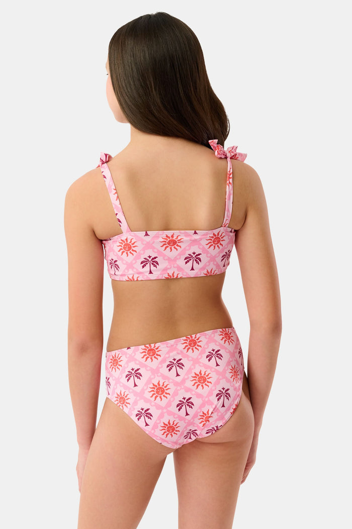 A young girl with long brown hair smiles in a Roller Rabbit Kids Sunny Days Cora Bikini, featuring sun and palm tree patterns and adjustable straps, against a plain light background.