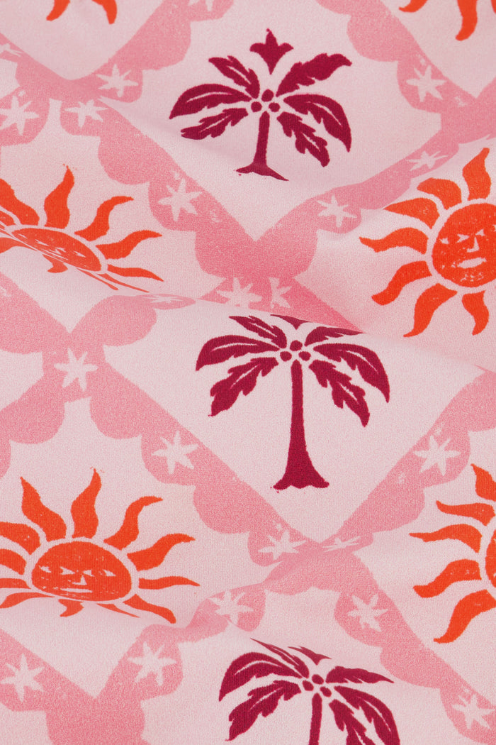 A young girl smiles in the Roller Rabbit Kids Sunny Days Rashguard—a pink long-sleeve one-piece swimsuit with orange and purple sun and palm tree prints—standing with one hand on her hip against a plain background.