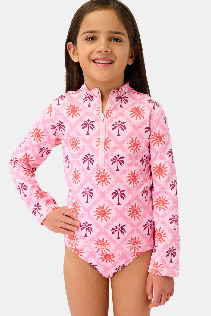 A young girl smiles in the Roller Rabbit Kids Sunny Days Rashguard—a pink long-sleeve one-piece swimsuit with orange and purple sun and palm tree prints—standing with one hand on her hip against a plain background.