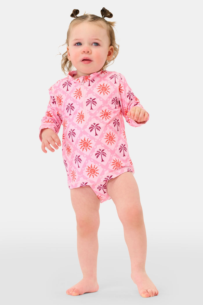 A young girl smiles in the Roller Rabbit Kids Sunny Days Rashguard—a pink long-sleeve one-piece swimsuit with orange and purple sun and palm tree prints—standing with one hand on her hip against a plain background.