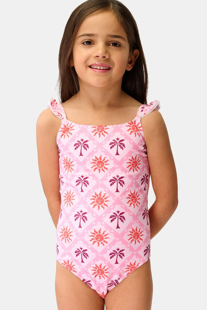 A young girl with long brown hair smiles in the Roller Rabbit Kids Sunny Days Rory One Piece, featuring ruffled straps and full coverage with orange and purple sun and palm tree patterns, against a plain light background.