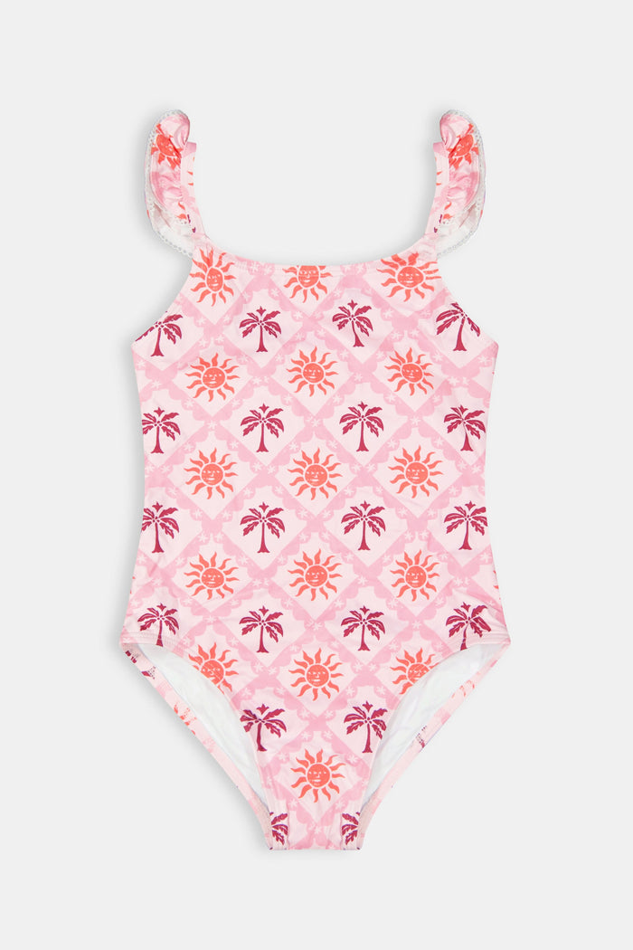 A young girl with long brown hair smiles in the Roller Rabbit Kids Sunny Days Rory One Piece, featuring ruffled straps and full coverage with orange and purple sun and palm tree patterns, against a plain light background.