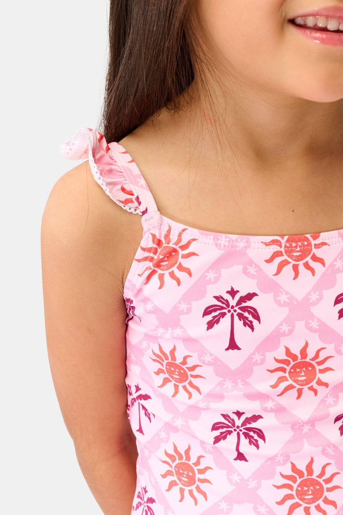 A young girl with long brown hair smiles in the Roller Rabbit Kids Sunny Days Rory One Piece, featuring ruffled straps and full coverage with orange and purple sun and palm tree patterns, against a plain light background.
