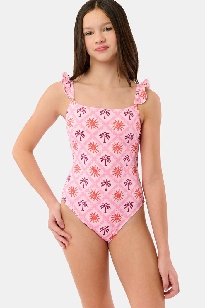 A young girl with long brown hair smiles in the Roller Rabbit Kids Sunny Days Rory One Piece, featuring ruffled straps and full coverage with orange and purple sun and palm tree patterns, against a plain light background.