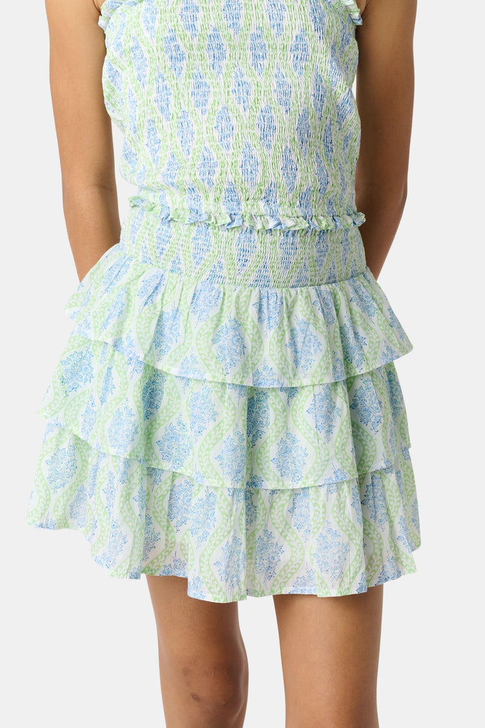A smiling girl wears the Roller Rabbit Kids Zelda Floral Sami Skirt as she stands by a blue bicycle near a marina, surrounded by others in summer outfits with boats, water, and houses in the background under a clear sky.