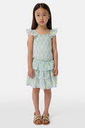 A smiling girl wears the Roller Rabbit Kids Zelda Floral Sami Skirt as she stands by a blue bicycle near a marina, surrounded by others in summer outfits with boats, water, and houses in the background under a clear sky.