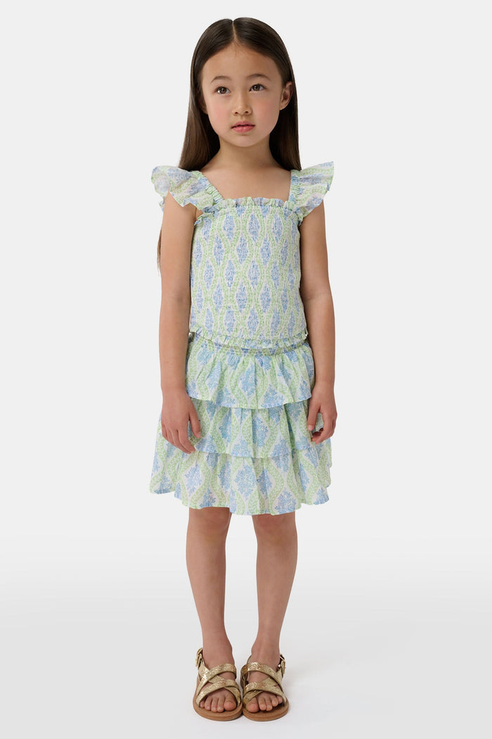 A smiling girl wears the Roller Rabbit Kids Zelda Floral Sami Skirt as she stands by a blue bicycle near a marina, surrounded by others in summer outfits with boats, water, and houses in the background under a clear sky.