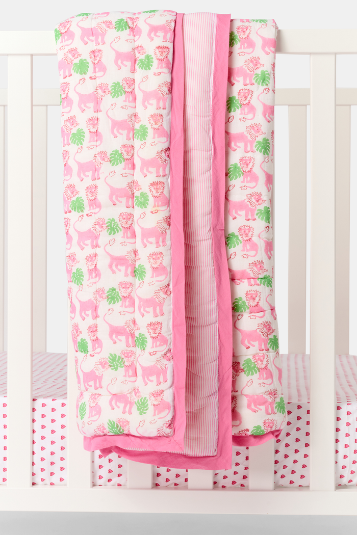 The Roller Rabbit Leo the Lion Baby Quilt features a soft cotton design with a pink border, white background, and playful pattern of pink elephants and green palm trees. A striped pink lining is visible along the folded edge.