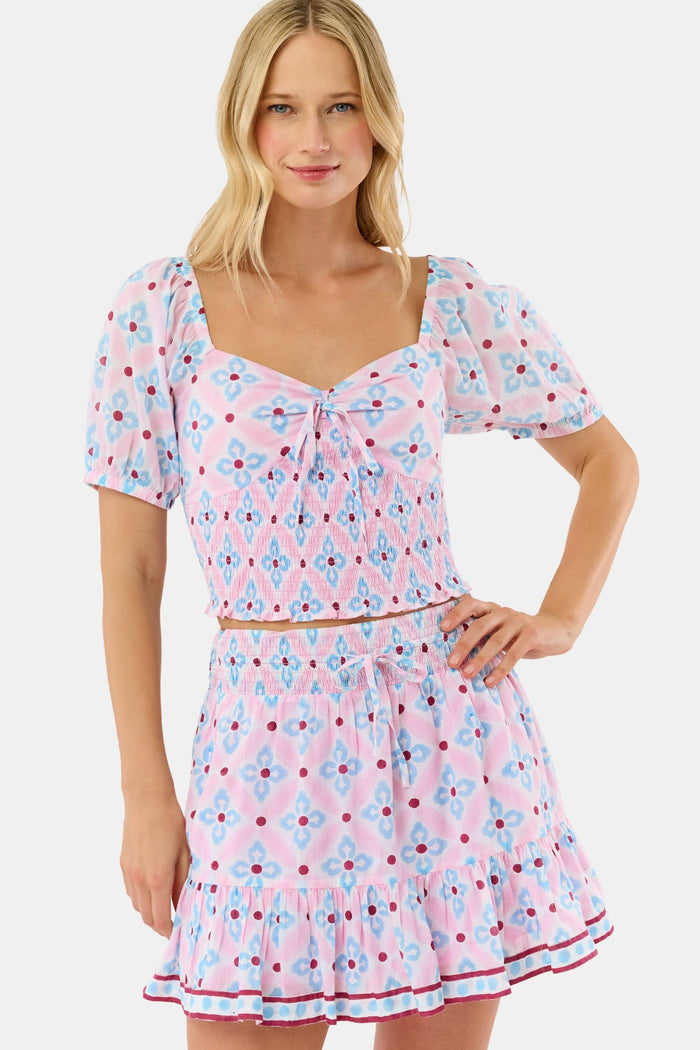 Wearing the Lerato Dovie Top by Roller Rabbit, a woman smiles with her hand on her hip. The light pink cotton crop top has a sweetheart neckline and features a blue and red floral geometric pattern.